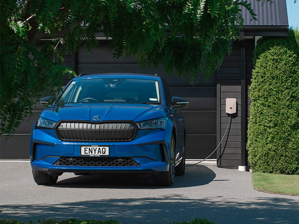 A blue enyaq plugged into an outdoor charger next to a residential garage